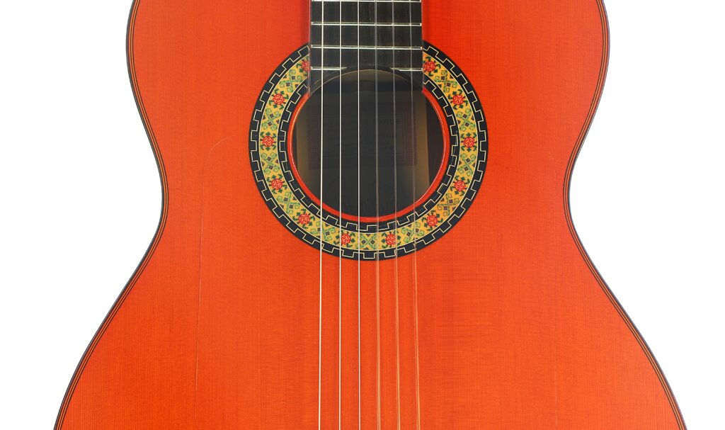 Eine Nahaufnahme des Korpus einer „Hermanos Conde 1998 Paco de Lucia Spezial Flamenco“-Gitarre, die sechs Saiten und eine kunstvoll gestaltete, farbenfrohe Rosette zeigt, die das Schallloch auf einem rötlichen Holzfinish umgibt. Eine Nahaufnahme des Korpus einer „Hermanos Conde 1998 Paco de Lucia Spezial Flamenco“-Gitarre, die sechs Saiten und eine kunstvoll gestaltete, farbenfrohe Rosette zeigt, die das Schallloch auf einem rötlichen Holzfinish umgibt.