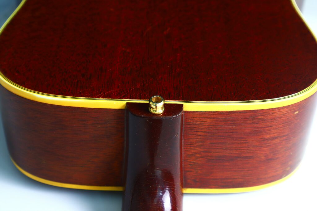 Nahaufnahme Des Rückens Einer Gibson Hummingbird 1966 Akustik Gitarre , Bei Der Der Korpus Auf Den Hals Trifft, Wobei Glänzendes Braunes Holz Mit Gelben Verzierungen Und Einem Metallischen Riemenknopf An Der Ferse Hervorgehoben Wird.