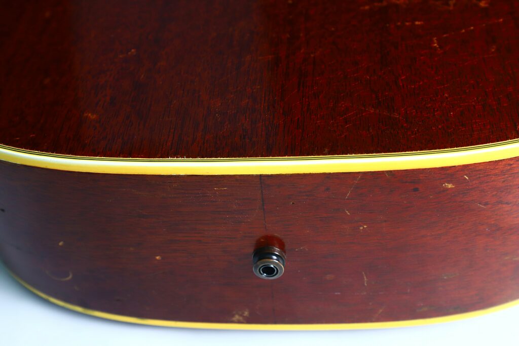 Nahaufnahme Der Seite Einer Gibson Hummingbird 1966 Akustik Gitarre Mit Glänzendem Holzkorpus, Gelber Verkleidung, Eingangsbuchse Und Sichtbaren Kratzern Und Gebrauchsspuren Auf Der Oberfläche.