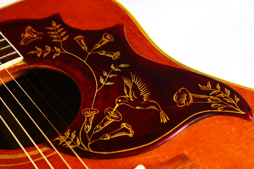 Nahaufnahme Des Schlagbretts Der Gibson Hummingbird 1966 Akustik Gitarre Mit Komplizierten Goldenen Blumen-, Blatt- Und Kolibri-Designs Auf Einem Rötlichen Holzhintergrund Mit Teilweise Sichtbaren Saiten Auf Der Linken Seite.