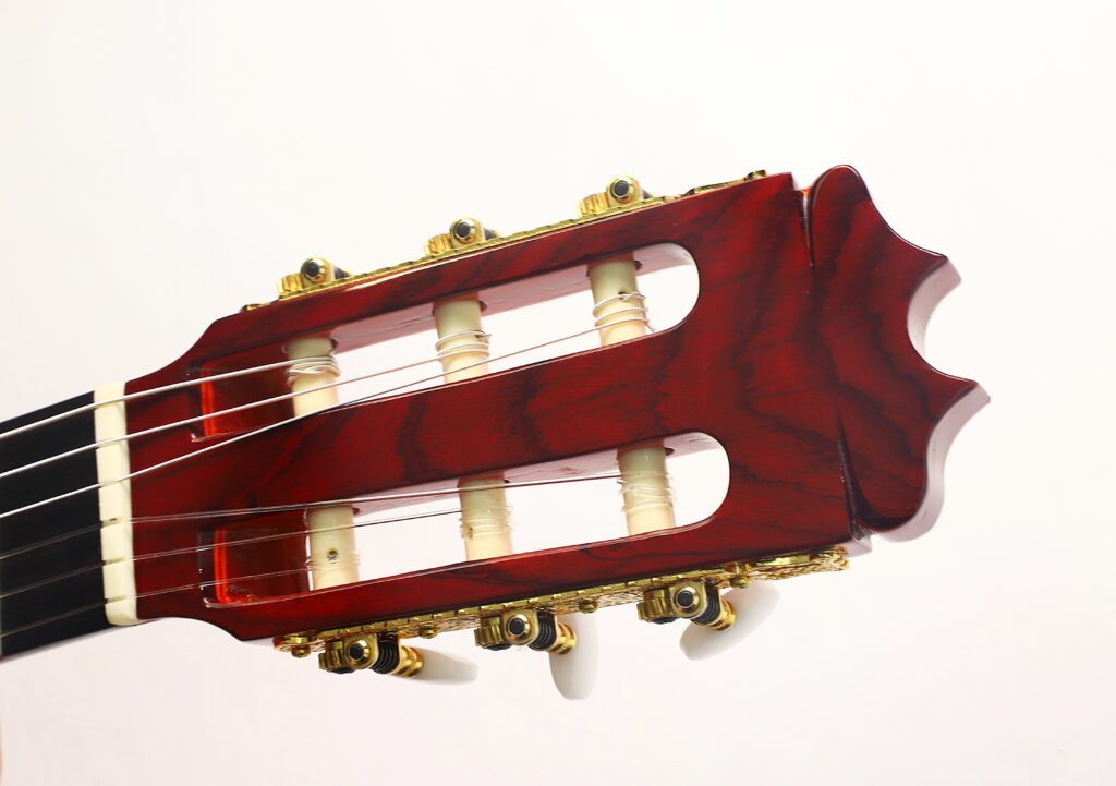 Nahaufnahme des Kopfstücks einer „Hermanos Conde 1998 Paco de Lucia Spezial Flamenco“-Gitarre, die goldene Stimmwirbel mit elfenbeinfarbenen Mechaniken und eine satte, rötliche Holzoberfläche vor weißem Hintergrund zeigt. Nahaufnahme des Kopfstücks einer „Hermanos Conde 1998 Paco de Lucia Spezial Flamenco“-Gitarre, die goldene Stimmwirbel mit elfenbeinfarbenen Mechaniken und eine satte, rötliche Holzoberfläche vor weißem Hintergrund zeigt.