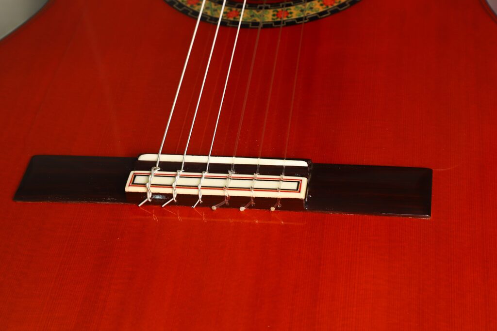 Nahaufnahme eines klassischen Gitarrenstegs, auf dem sechs Nylonsaiten zu sehen sind, die am Holzkorpus einer „Hermanos Conde 1998 Paco de Lucia Spezial Flamenco-Gitarre“ befestigt sind; oben ist teilweise eine dekorative Schalllochrosette zu erkennen. Nahaufnahme eines klassischen Gitarrenstegs, auf dem sechs Nylonsaiten zu sehen sind, die am Holzkorpus einer „Hermanos Conde 1998 Paco de Lucia Spezial Flamenco-Gitarre“ befestigt sind; oben ist teilweise eine dekorative Schalllochrosette zu erkennen.