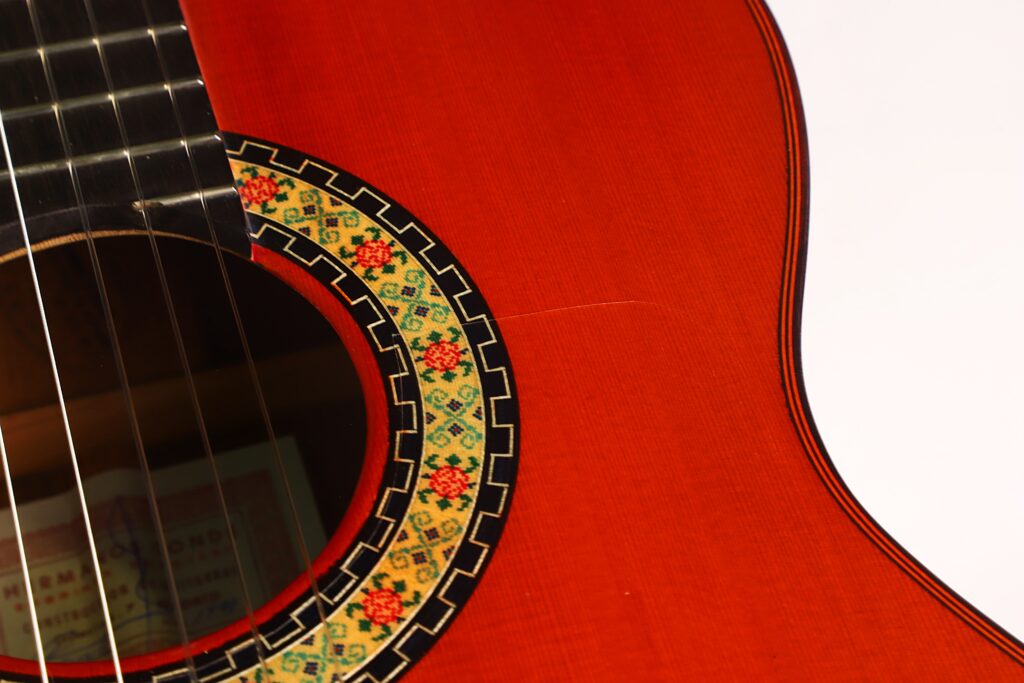 Nahaufnahme des Schalllochs einer „Hermanos Conde 1998 Paco de Lucia Spezial Flamenco“-Gitarre mit einem detailreichen, farbenfrohen Mosaik-Rosettenmuster, einem Ausschnitt des Griffbretts und einem rotbraunen Holzkorpus. Rechts ist ein weißer Hintergrund zu sehen. Nahaufnahme des Schalllochs einer „Hermanos Conde 1998 Paco de Lucia Spezial Flamenco“-Gitarre mit einem detailreichen, farbenfrohen Mosaik-Rosettenmuster, einem Ausschnitt des Griffbretts und einem rotbraunen Holzkorpus. Rechts ist ein weißer Hintergrund zu sehen.
