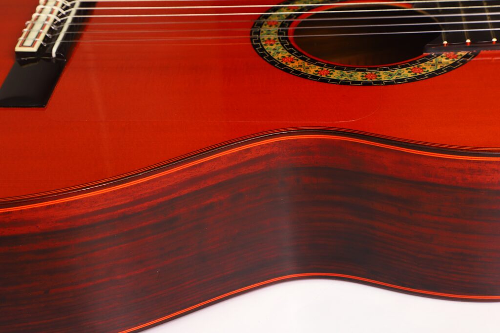 Nahaufnahme des Korpus einer „Hermanos Conde 1998 Paco de Lucia Spezial Flamenco“-Gitarre, die die satte, rötliche Holzmaserung, die filigrane Rosette um das Schallloch sowie Teile der Saiten und des Stegs vor weißem Hintergrund hervorhebt. Nahaufnahme des Korpus einer „Hermanos Conde 1998 Paco de Lucia Spezial Flamenco“-Gitarre, die die satte, rötliche Holzmaserung, die filigrane Rosette um das Schallloch sowie Teile der Saiten und des Stegs vor weißem Hintergrund hervorhebt.