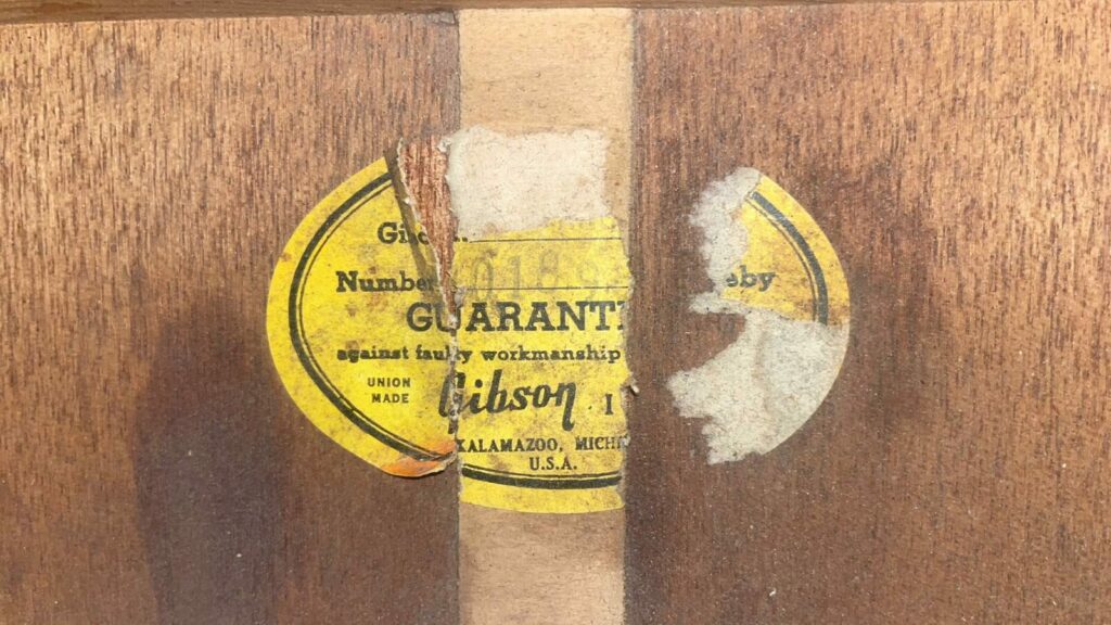 Auf Dem Holz Einer Gibson Hummingbird-Akustikgitarre Von 1966 Ist Ein Teilweise Zerrissenes Gelbes Gibson-Etikett Angebracht, Auf Dem Garantiert Gegen Verarbeitungsfehler Und Kalamazoo, Michigan, U.s.a. Mit Verblasstem Oder Fehlendem Text Steht.