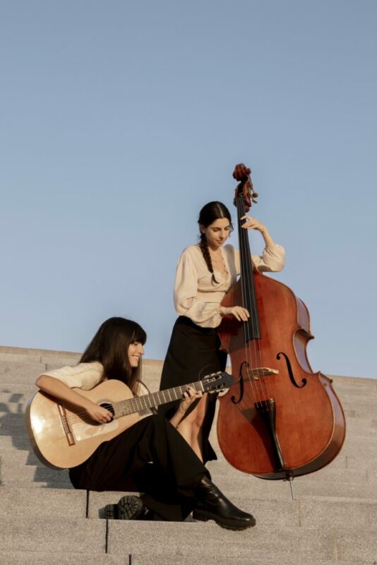 Two women perform music outdoors on stone steps; one is seated playing an acoustic guitar, while the other stands playing a double bass. Both wear light blouses and dark skirts under a clear blue sky.