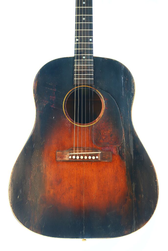 A Vintage Gibson J-45 1951 Acoustic Guitar With A Sunburst Finish, Visible Scratches, And Darkened Edges Is Displayed Against A Plain White Background.