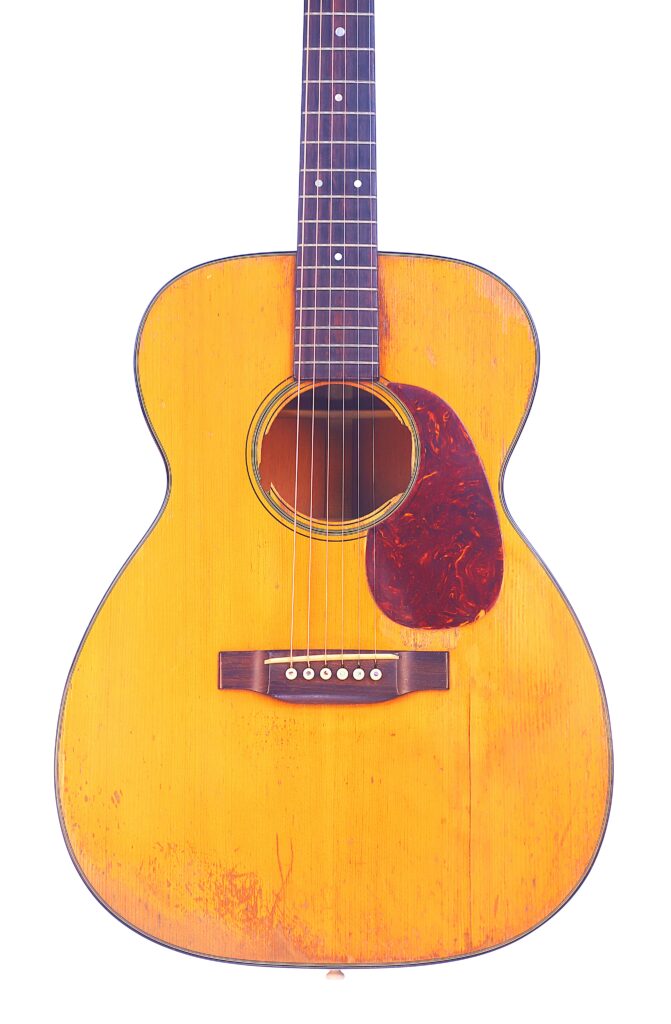 A Martin 00-18 1952 Acoustic Guitar Featuring A Worn, Yellowish-Brown Wooden Body, Six Strings, And A Dark Tortoiseshell Pickguard Is Shown Against A White Background.