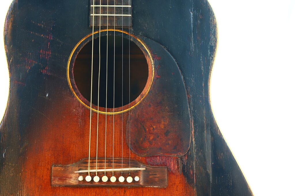 Close-Up Of A Gibson J-45 1951 Acoustic Guitar With A Weathered Wooden Body, Visible Scratches, Faded Areas, Strings, And Pickguard Against A White Background—Signs Of Frequent Use.