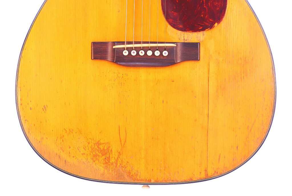 Close-Up Of The Martin 00-18 1952 Acoustic Guitar’s Lower Half, Highlighting Its Wooden Bridge, Six Strings, Light Brown Worn Surface With Visible Scratches, And Dark Tortoiseshell Pickguard.