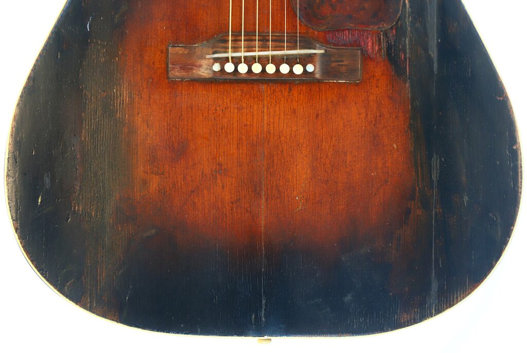 Close-Up Of The Lower Half Of A Gibson J-45 1951 Acoustic Guitar, Featuring A Worn Wooden Surface With Scratches, Six Strings, Bridge, White Bridge Pins, And A Finish That Fades From Dark Edges To A Lighter Center.