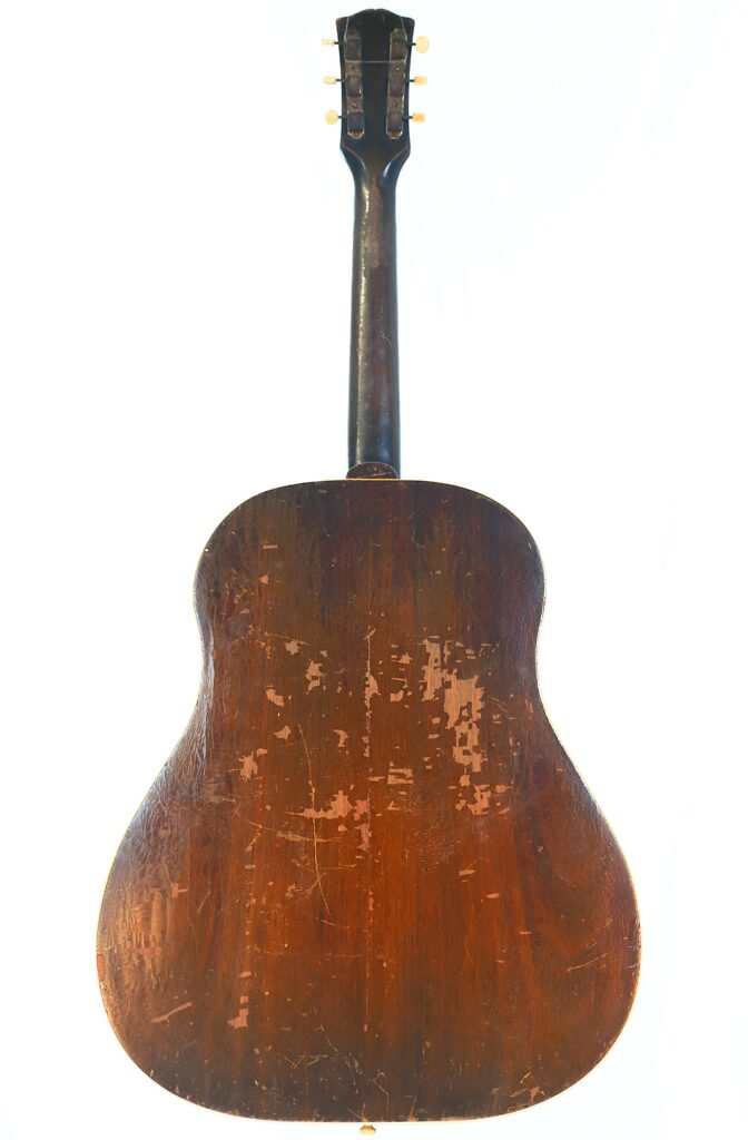 The Back Of A Well-Worn Gibson J-45 1951 Acoustic Guitar Shows Its Dark Wood Finish With Many Scratches, Scuffs, And Signs Of Age. The Tuning Pegs Are Visible At The Top Of The Neck.