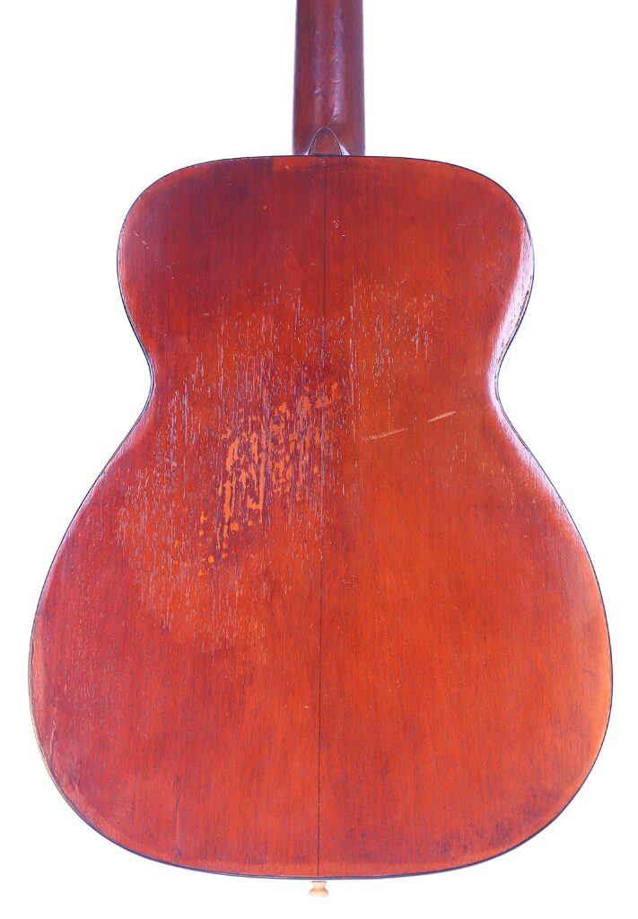 The Image Shows The Back Of A Martin 00-18 1952 Acoustic Guitar With A Wooden Finish, Visible Wear Marks, And A Simple, Classic Shape. The Wood Appears Slightly Scratched And Aged, Highlighting Its Vintage Character.