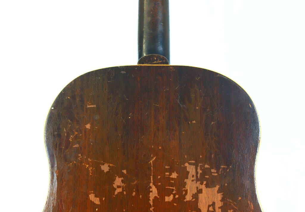 Close-Up Of The Back Of A Vintage Gibson J-45 1951 Acoustic Guitar, Showing Visible Scratches And Faded Wood Areas, Photographed Against A White Background.