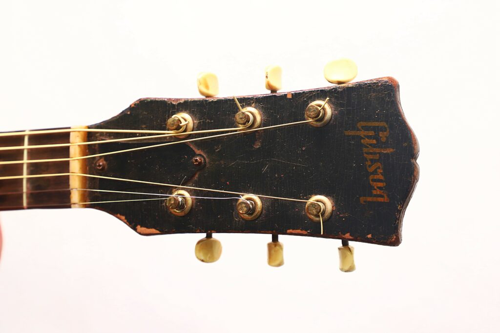 Close-Up Of A Worn Gibson J-45 1951 Acoustic Guitar Headstock With Yellow Tuning Pegs And A Partially Faded Logo, Showing Signs Of Use And Age Against A Plain White Background.