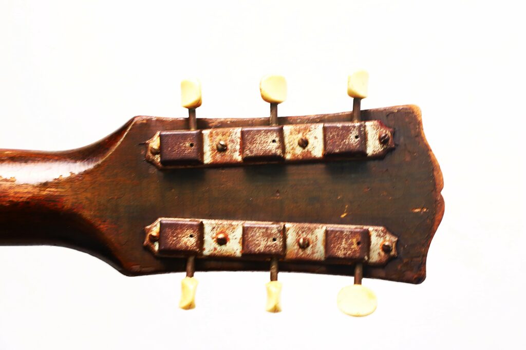 Close-Up Of The Back Of A Worn Gibson J-45 1951 Acoustic Guitar Headstock With Two Metal Tuning Machine Strips And Cream-Colored Pegs, Set Against A Plain White Background.
