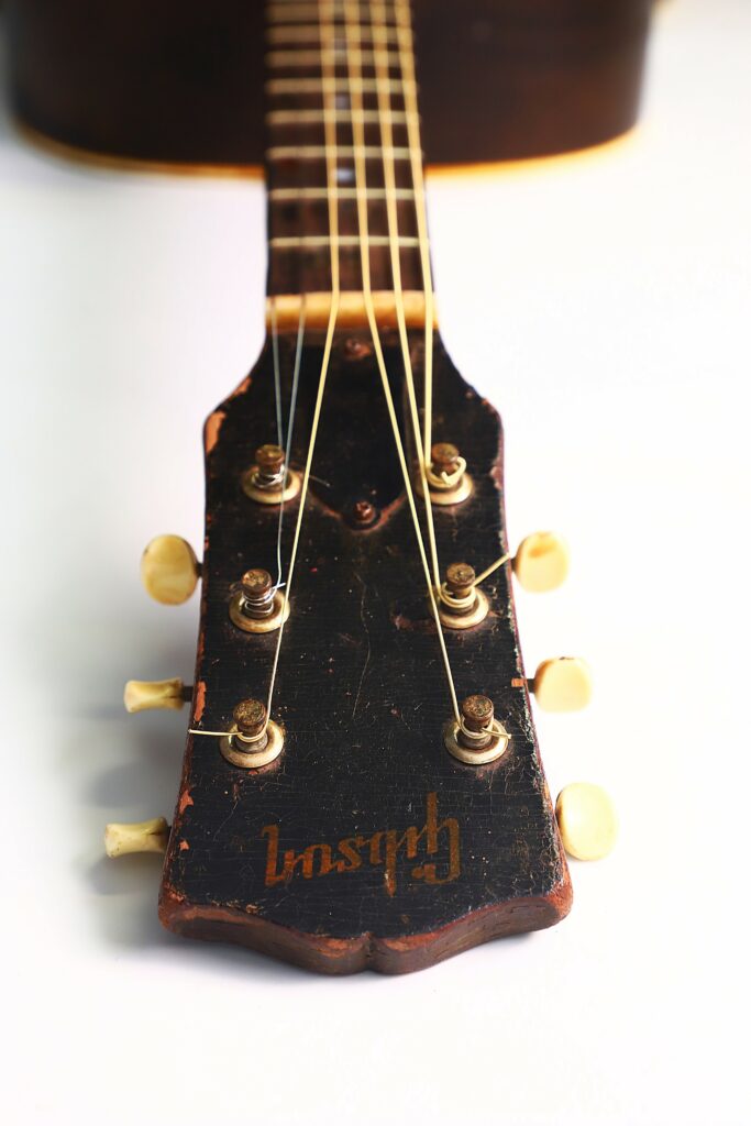 Close-Up Of A Vintage Gibson J-45 1951 Acoustic Guitar Headstock With Six Tuning Pegs And Visible Strings, Photographed Against A White Background. The Gibson Logo Appears Upside Down.