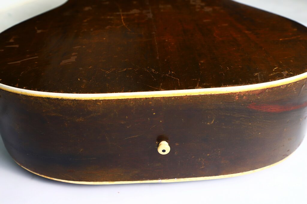 Close-Up Of The Side And Back Of A Gibson J-45 1951 Acoustic Guitar, Showing Worn, Dark Wood With Visible Scratches, A White Strap Button In The Center, And Light-Colored Binding Along The Edges.