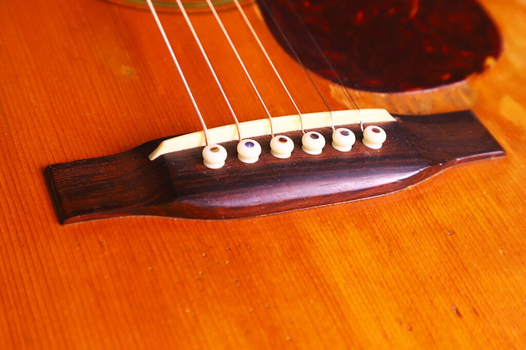 Close-Up Of The Bridge And Strings Of A Martin 00-18 1953 Acoustic Guitar On A Wooden Surface, Highlighting The White Bridge Pins And Part Of The Pickguard.