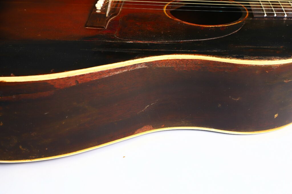 Close-Up Of The Side And Body Of A Worn Gibson J-45 1951 Acoustic Guitar With Dark Brown Finish, Yellowed Trim, And Visible Scratches, Highlighting Signs Of Age Against A White Background.