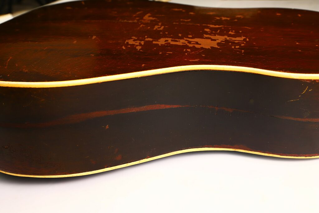 Close-Up Of The Side And Back Of A Worn Gibson J-45 1951 Acoustic Guitar With A Dark Brown Finish, Visible Scratches, And Cream-Colored Binding Along The Edges, Resting On A White Surface.