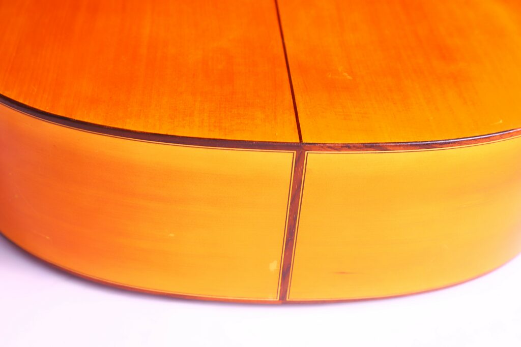 Close-Up Of The Bottom Edge Of A Jose Ramirez 1968 Flamenco Guitar, Highlighting Its Smooth, Shiny Wooden Surface And The Seam Where Two Wood Pieces Meet. The Image Is Brightly Lit Against A White Background.