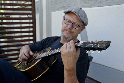 A Man Wearing Glasses And A Gray Flat Cap Is Sitting Outdoors, Playing An Acoustic Guitar And Looking Focused.