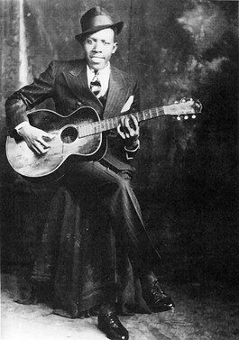 Robert Johnson: The Legendary Delta Bluesman - Black-And-White Photo Of A Man In A Suit, Tie, And Hat Sitting On A Stool, Playing An Acoustic Guitar And Smiling At The Camera. The Background Is Dark And Plain.