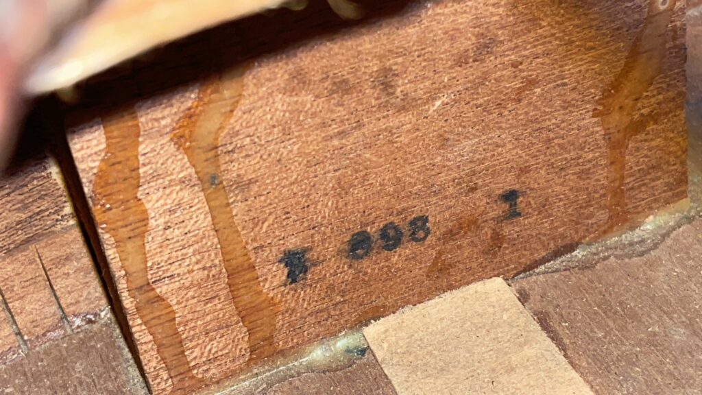 Close-Up Of A Gibson J-45 1951 Acoustic Guitar Wooden Surface, Showing The Black-Stamped Marking 7 898 I Along With Visible Wood Grain And Glue Residue.
