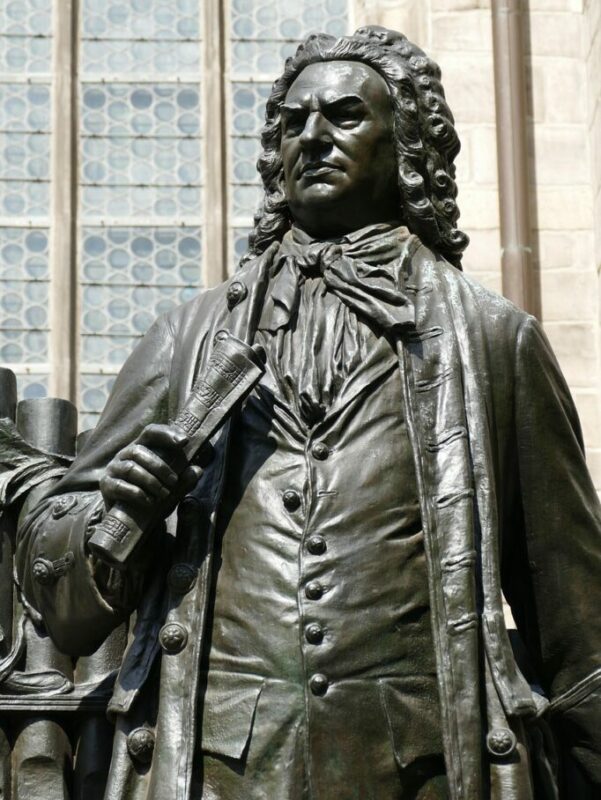 A Bronze Statue Of A Man In Historical Clothing With Curly Hair, Holding A Rolled-Up Document, Stands In Front Of A Building With Large, Patterned Windows.