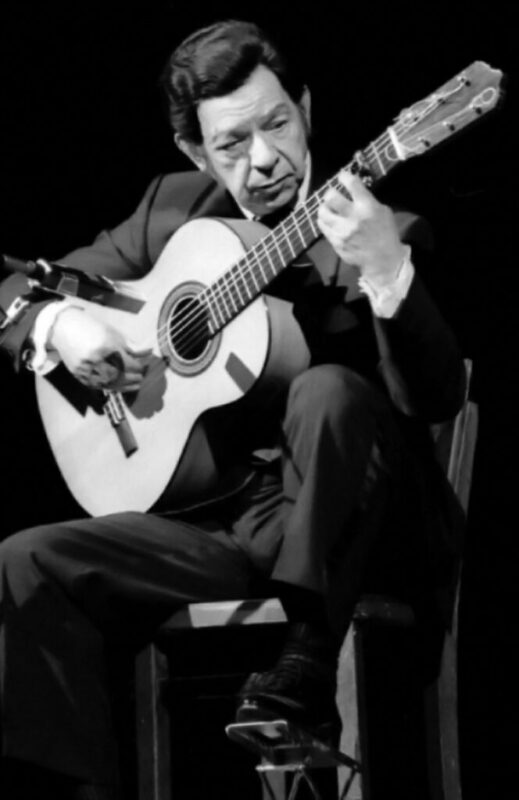 A Man In A Suit Sits On A Chair, Playing A Classical Guitar. He Appears Focused On His Performance, With One Leg Crossed Over The Other, And Is Positioned Against A Dark Background.