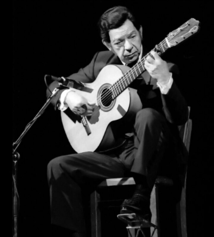 Sabicas - A Man In A Suit Sits On A Chair Playing A Classical Guitar On Stage, Deeply Focused, With A Microphone Positioned In Front Of Him. The Background Is Dark, Highlighting The Performer.