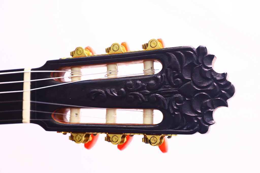Close-Up Of A José Ruiz Pedregosa 2020 Classical Guitar Headstock Featuring Ornate Carving, Gold Tuning Pegs, And Orange Tuning Knobs Against A White Background.