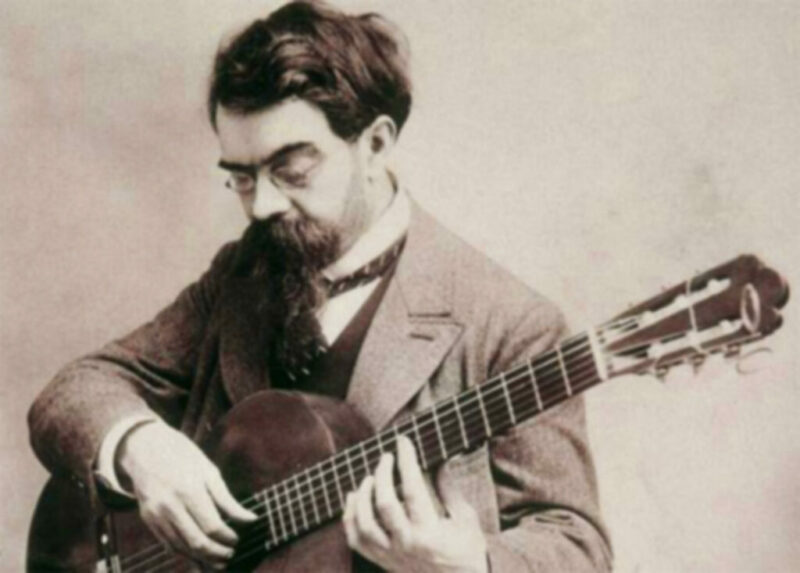 A Bearded Man Wearing Glasses And A Suit Plays A Classical Guitar, Focusing Intently On The Strings With Both Hands On The Instrument. The Image Is In Black And White.