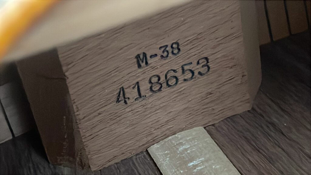 A Close-Up Of A Martin M-38 1979 Acoustic Guitar’s Wooden Block, Marked “M-38” And Numbered “4,18653” In Black Ink, Rests On A Wooden Surface With Visible Nearby Planks.