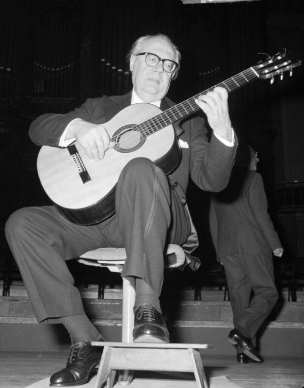 Andrés Segovia - The Legendary Classical Guitarist - Playing A Classical Guitar On Stage In The Style Of Andrés Segovia. Another Person In A Suit Walks In The Background, Suggesting The Scene Is During A Rehearsal Or Performance.