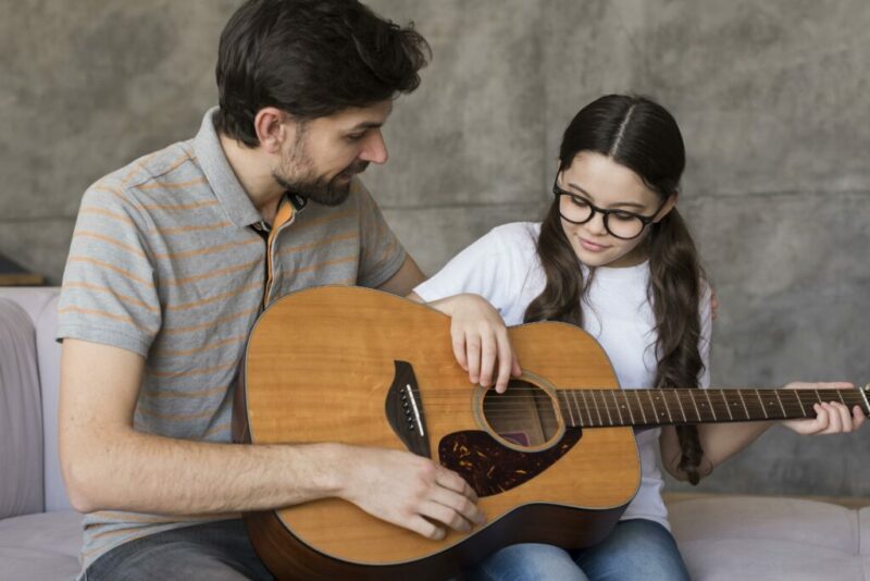 A Man Teaches A Young Girl How To Play An Acoustic Guitar. They Sit Together On A Couch, With The Man Guiding The Girls Hands On The Strings. The Girl Wears Glasses And Both Appear Focused And Engaged.