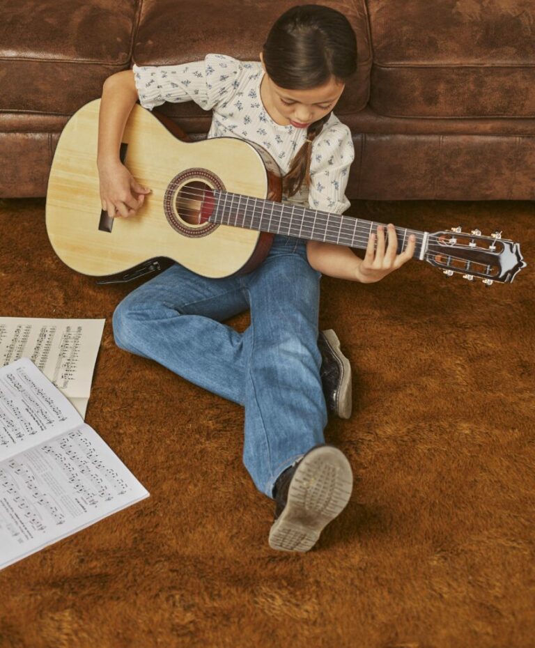 Gitarre Lernen - Ein junges Mädchen sitzt auf einem braunen Teppich und spielt auf einer Akustikgitarre, vor der sie Notenblätter ausgebreitet hat. Sie ist auf ihr Spiel konzentriert und trägt ein weißes gemustertes Oberteil und blaue Jeans.
