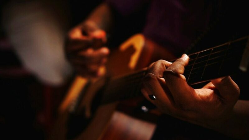 Fingerstyle Guitar -Close-Up Of A Persons Hands Playing An Acoustic Guitar, With Fingers Pressing The Strings On The Fretboard. The Background Is Dark And Out Of Focus