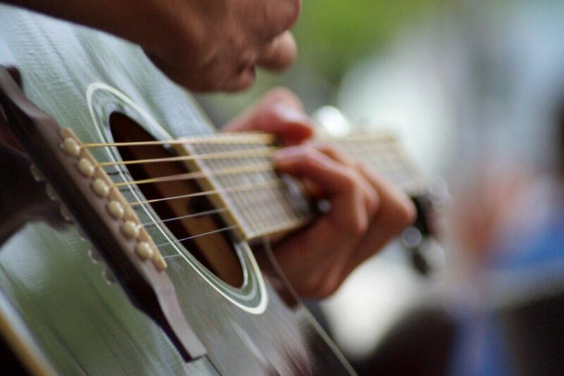 Close-Up Of A Person’s Hands Playing An Acoustic Guitar, With Fingers Pressing The Strings On The Fretboard And The Other Hand Strumming Near The Sound Hole. The Background Is Blurred.