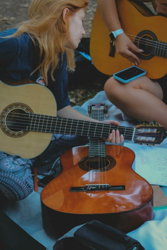 Two People Sit On A Blanket Outdoors, Each Holding An Acoustic Guitar. One Person Looks To The Side While The Other Strums. A Phone And Sheet Music Are Placed Nearby.