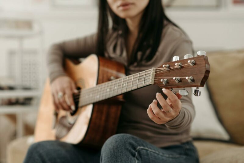 Guitar Tuning - A Person Sitting On A Couch Playing An Acoustic Guitar, Focusing On Tuning The Instrument. The Person Wears A Long-Sleeved Shirt And Jeans, With Their Face Partially Out Of Frame.
