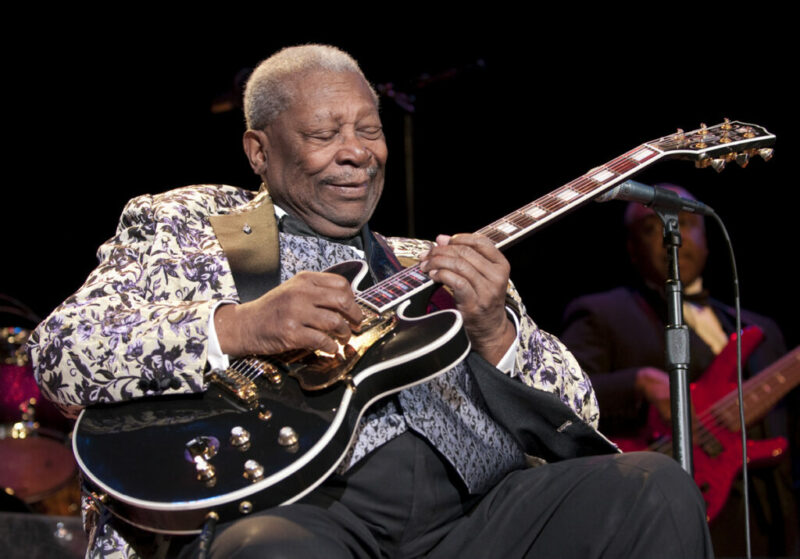 An Older Man In A Patterned Jacket Sits And Plays An Electric Guitar On Stage, Eyes Closed And Smiling, With Another Musician And Musical Instruments Visible In The Background.