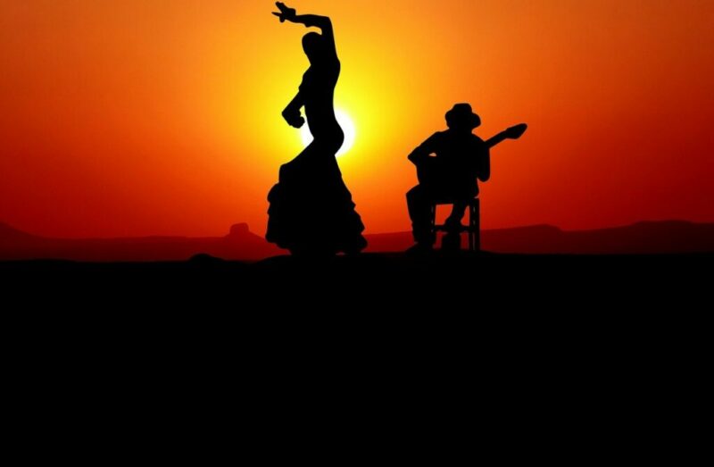 The History Of Flamenco Guitar - Silhouette Of A Flamenco Dancer With One Arm Raised And A Seated Guitarist Playing, Set Against A Vivid Orange Sunset