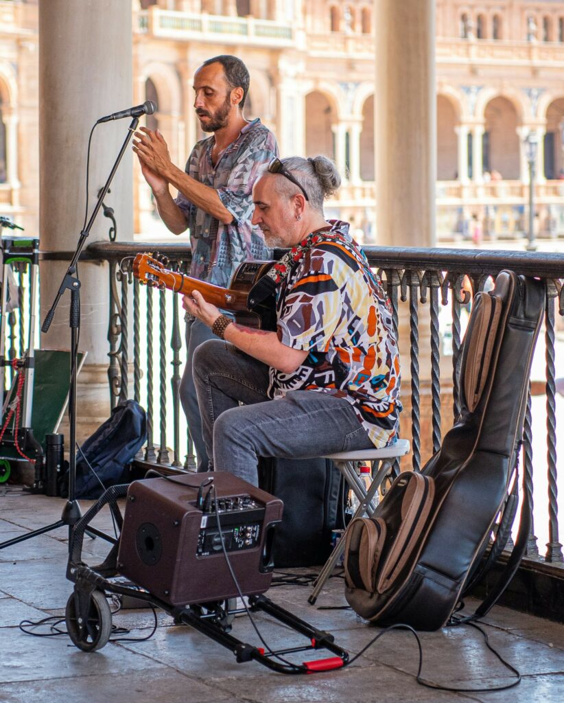 Zwei Männer musizieren im Freien; einer spielt im Sitzen eine akustische Gitarre, der andere singt in ein Mikrofon. In der Nähe befinden sich Musikgeräte und ein Gitarrenkoffer. Im Hintergrund ist ein Gebäude mit Bögen zu sehen.