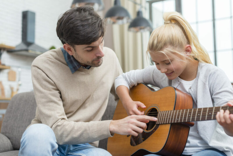 A Man Is Teaching A Smiling Young Girl To Play An Acoustic Guitar, Guiding Her Fingers On The Strings As They Sit Together On A Couch In A Cozy, Well-Lit Room.
