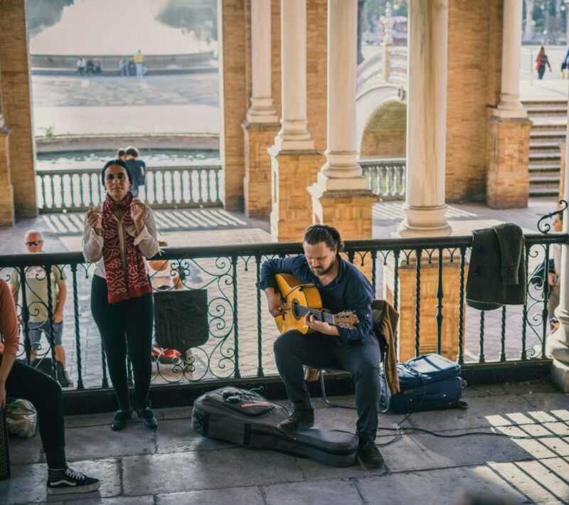 A Man Sits And Plays Guitar While A Woman Stands Clapping Beside Him Under An Ornate Covered Area, With A Fountain, Bridge, And People Visible In The Sunny Background.