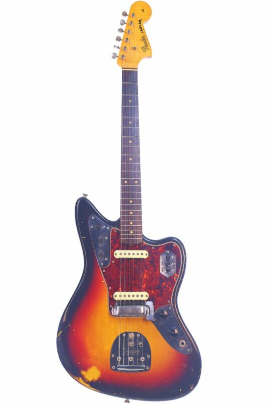 Die Fender Jaguar 1963 E-Gitarre verfügt über ein Sunburst-Finish, abgenutzte Kanten, zwei Tonabnehmer, ein Schildpatt-Schlagbrett und einen Ahorn-Hals mit dem Fender-Logo auf der Kopfplatte.