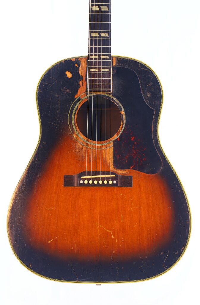 The Gibson Southern Jumbo 1955 Acoustic Guitar Features A Sunburst Finish, Six Strings, And A Pickguard. Visible Body Wear, Including Scratches And Worn Spots, Indicates The Instrument Has Seen Frequent Use Over Time.