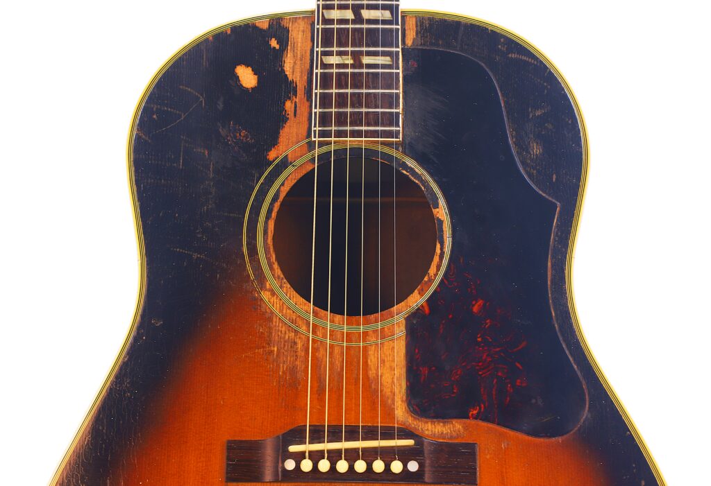 Close-Up Of A Gibson Southern Jumbo 1955 Acoustic Guitar In Sunburst Finish, Showing Visible Scratches And Chipped Paint Near The Sound Hole And Pickguard, Set Against A White Background.